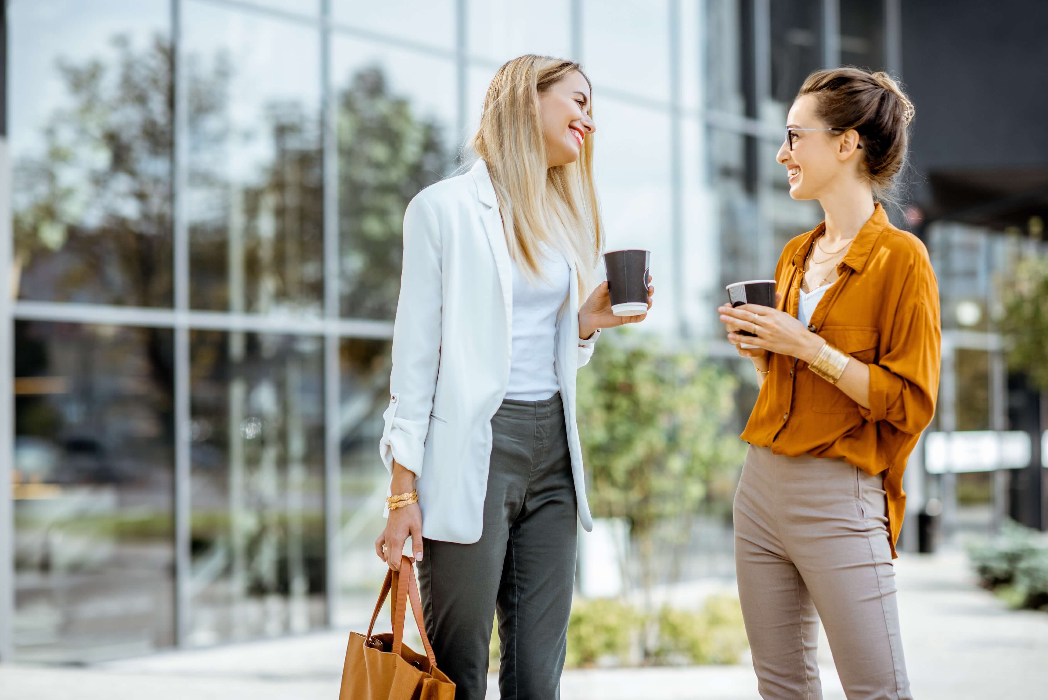 Two women talking outside, smiling and holding coffee.
