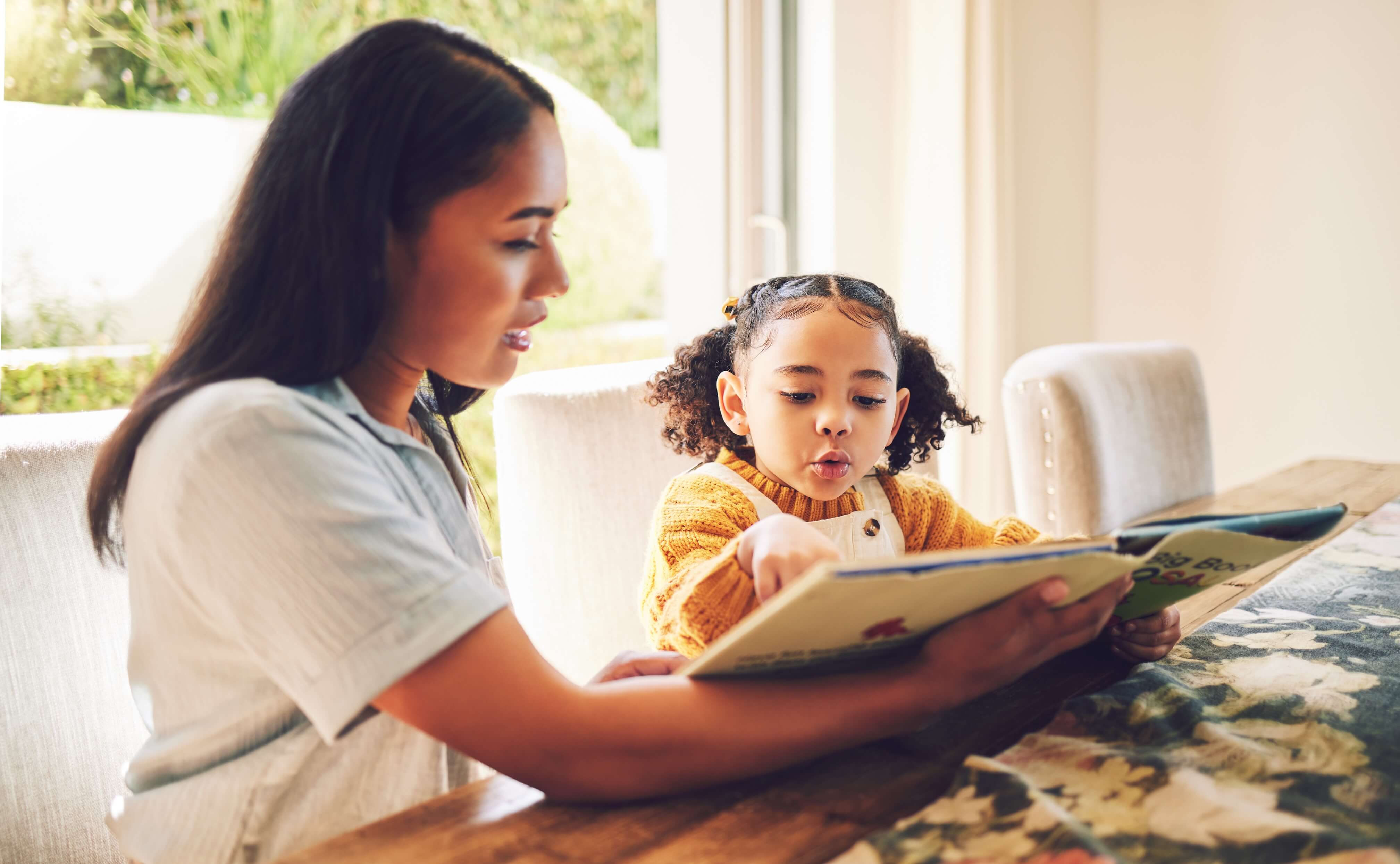Woman is reading a book with her child while seating at a table.