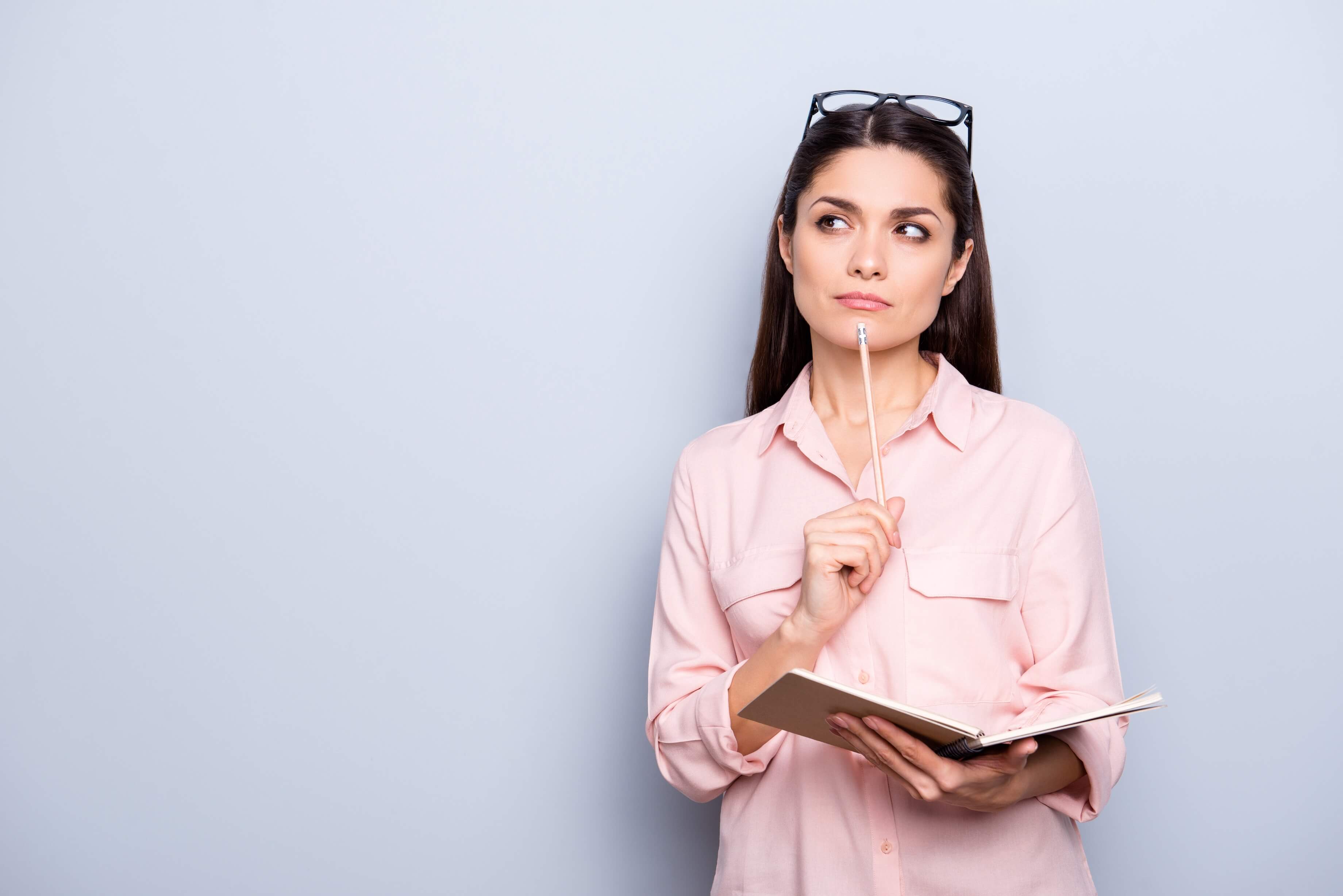 Woman in a pink shirt stands holding a book and a pencil while thinking.
